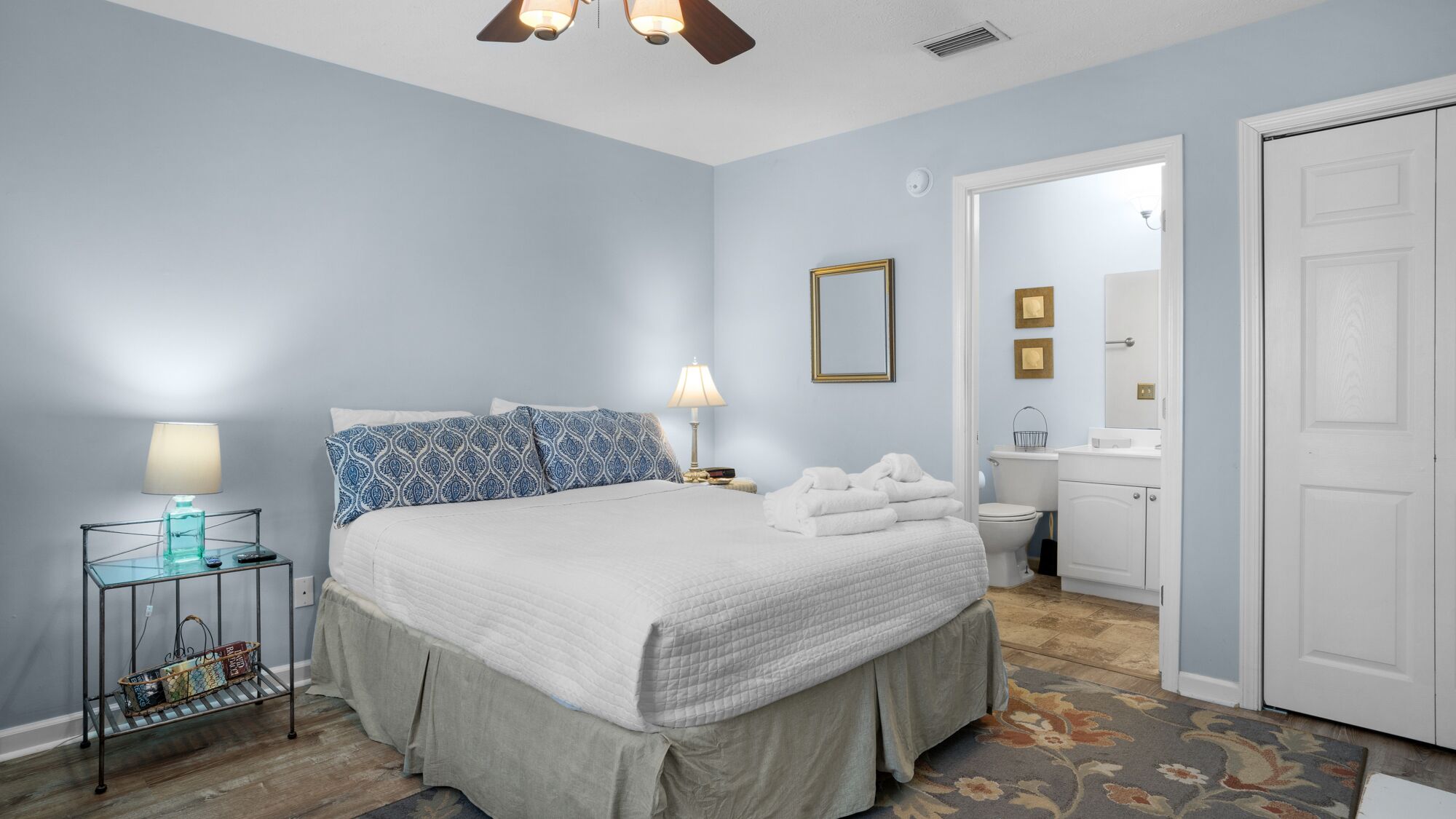 Un dormitorio con cama, mesitas de noche, lámpara, espejo y puerta que da a un baño.