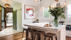 A modern kitchen with a white island, bar stools, a large window, and a decorative plant. - Stockton House- light-filled retreat with art and unique flair (Daylesford)