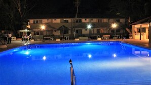 A swimming pool at night with surrounding buildings and a few people.