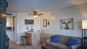 A living room with a sofa, a coffee table, and a kitchen area with a bar stool. - Cozy Condo with Views of Mt Kineo (Rockwood)