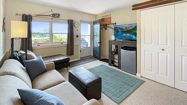 A living room with a sofa, a television, a small refrigerator, and a window with a view of a car and greenery outside. - Cozy Condo with Views of Mt Kineo (Rockwood)