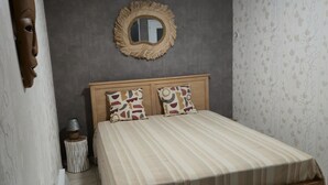 Un dormitorio con una cama de madera, dos almohadas estampadas, un espejo redondo y una mesita de noche con una lámpara.