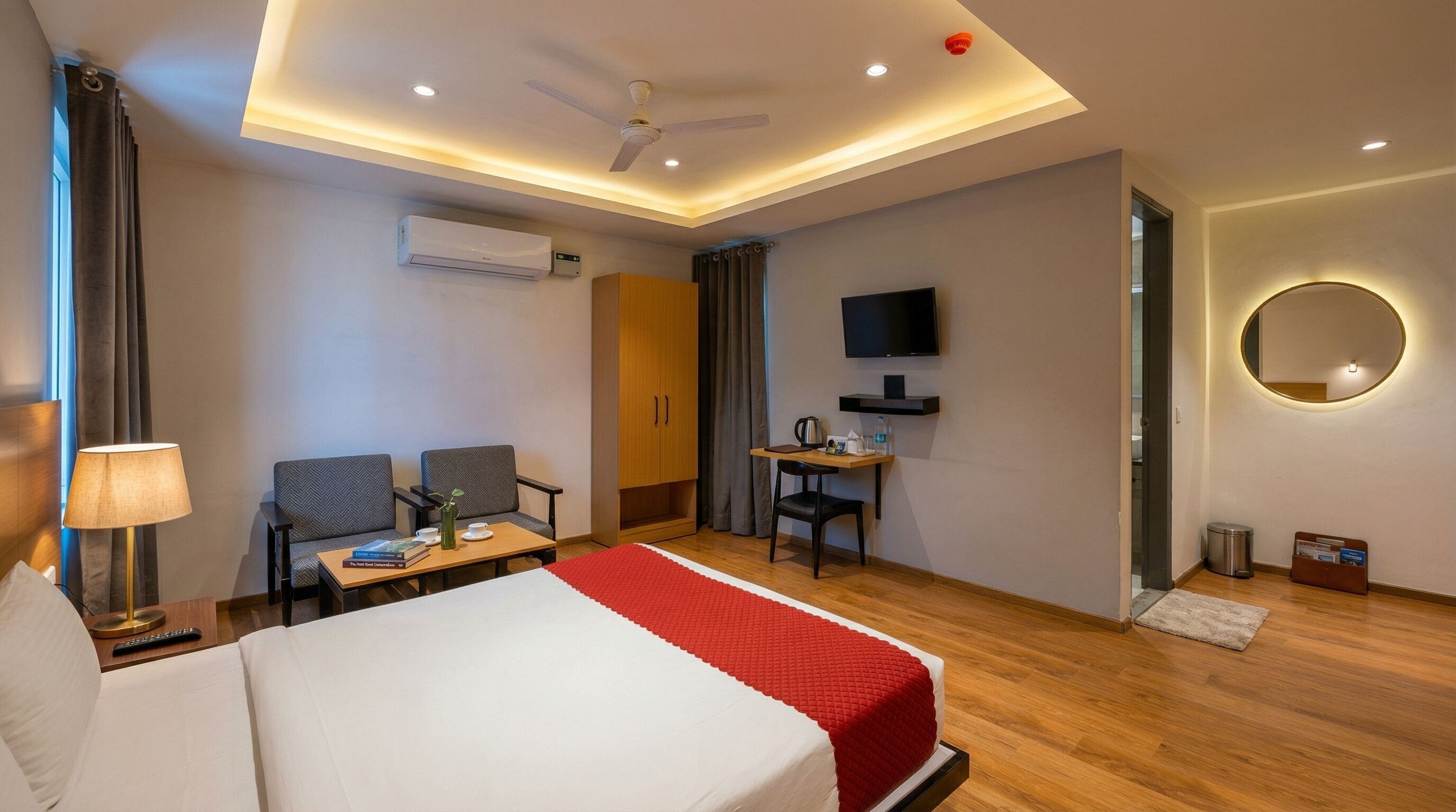 A hotel room with a bed, a desk, two chairs, a TV, a ceiling fan, an air conditioner, and a mirror.