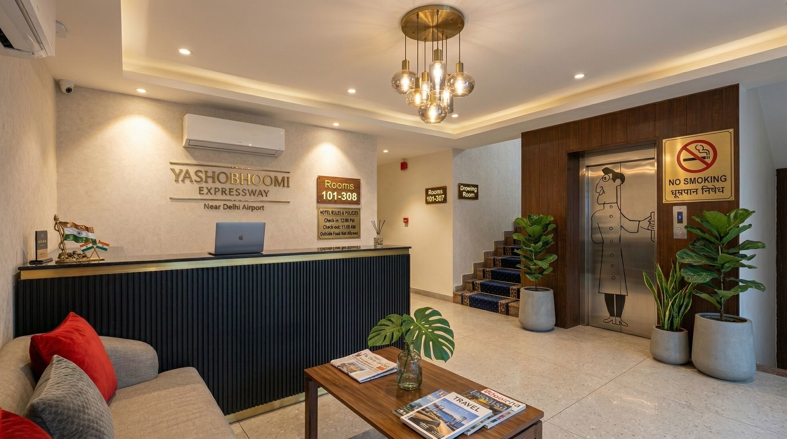 A hotel reception area with a reception desk, a laptop, a coffee table with magazines, a staircase, and a no-smoking sign.