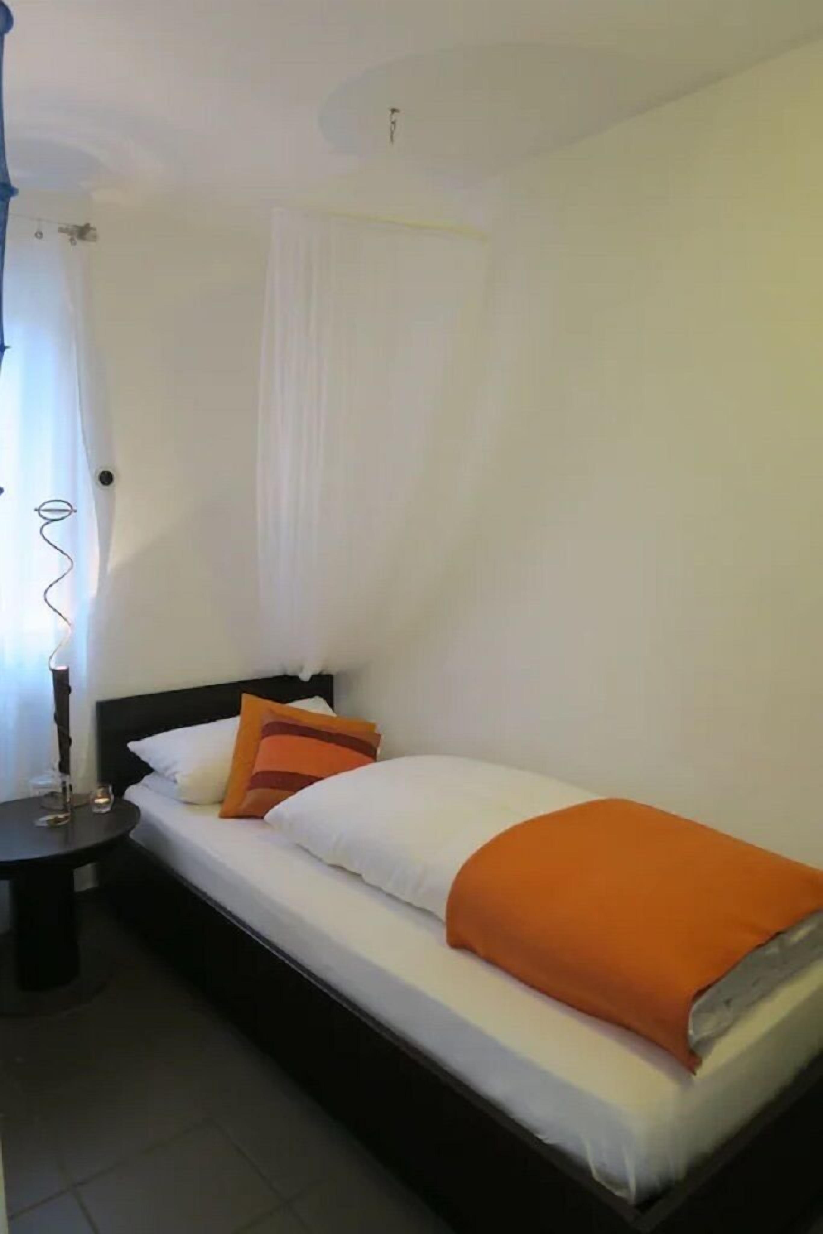 Een kleine kamer met een enkel bed, witte wanden en een donker hoofdeinde. Er staat een nachtkastje met een lamp en een tafeltje met een glas.