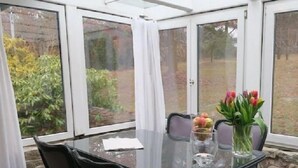 A conservatory with a glass table and chairs, a vase with flowers, and a view of the garden outside. - Seagull house, beach, bathing jetty, boat mooring on Lake Plau (Alt Schwerin)