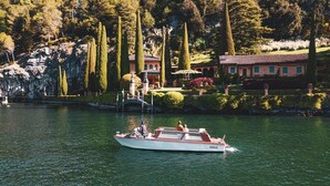 A boat on a calm lake with a rocky shoreline and a building with a red roof in the background. - Il Cuore del Lago in Varenna (Varenna)