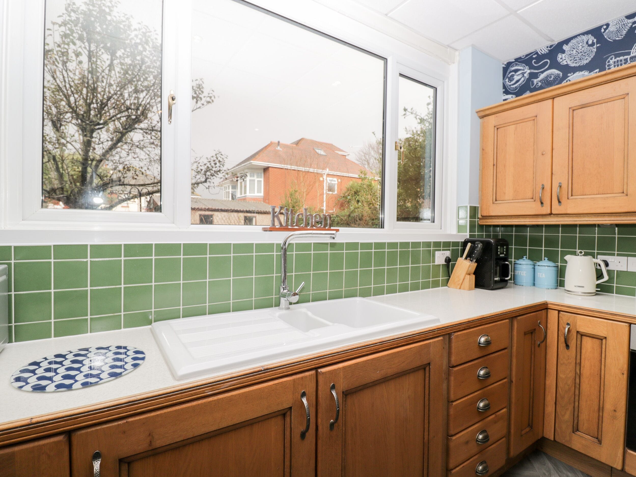 Una cocina con un ventanal grande, azulejos verdes, armarios de madera y un lavamanos.