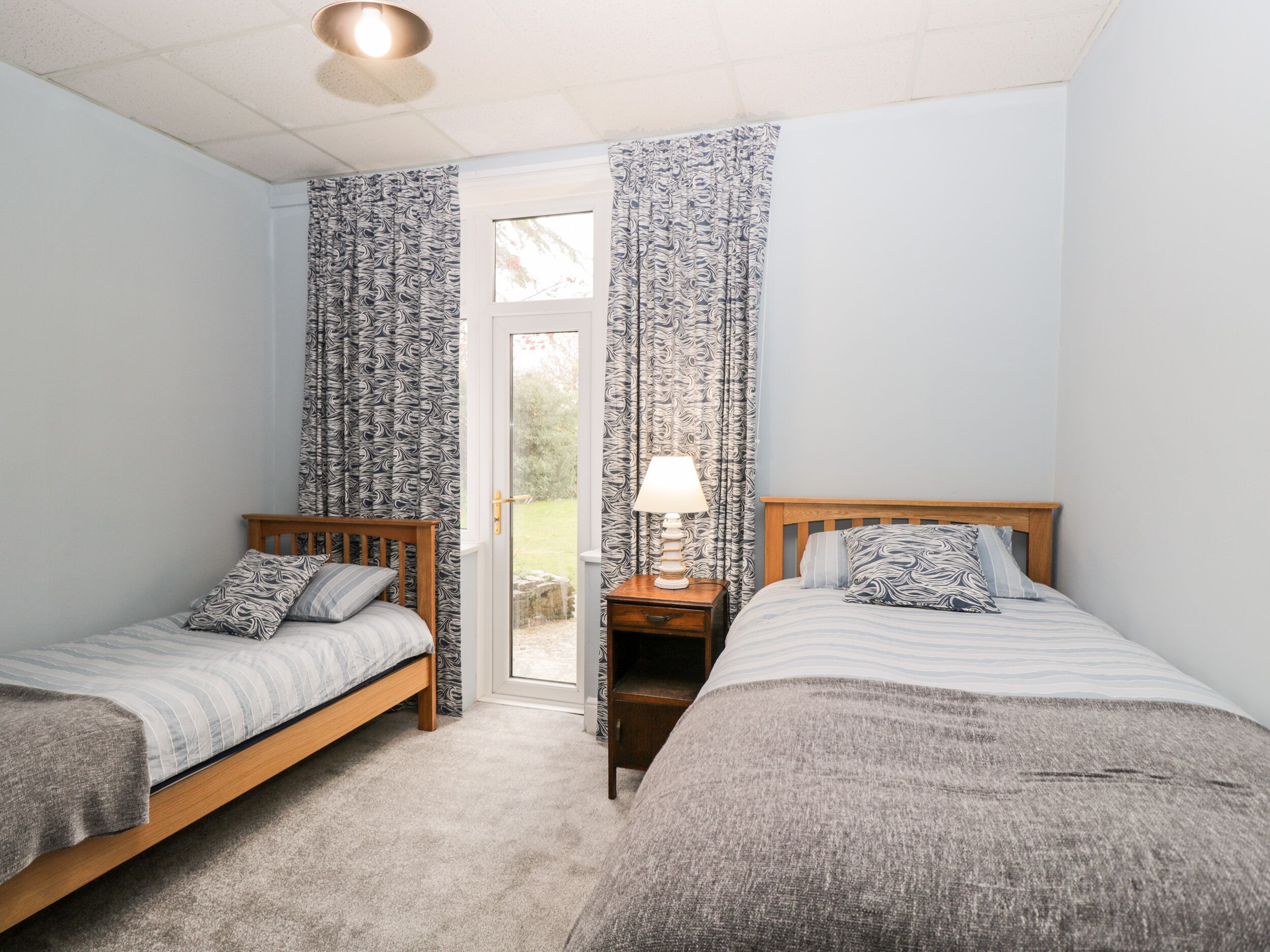 A bedroom with two single beds, a nightstand with a lamp, and a window with patterned curtains.