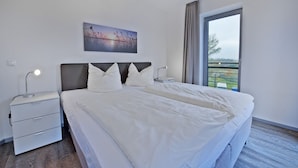 Ein modernes Hotelzimmer mit einem großen Bett, zwei Nachttischen mit Lampen, einem Bild an der Wand und einem großen Fenster mit Blick ins Grüne.