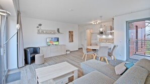 Ein modernes Wohnzimmer mit einer Couch, einem Couchtisch und einem Fernseher. Es gibt einen Essbereich mit Tisch und Stühlen. Das Zimmer hat ein großes Fenster und blickt auf ein Backsteinhaus.