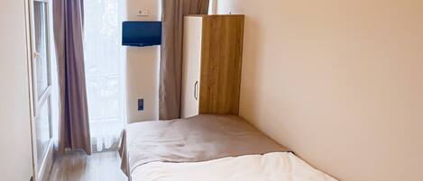 A small, single-bed room with a wooden wardrobe, a ceiling fan, and a window with curtains.
