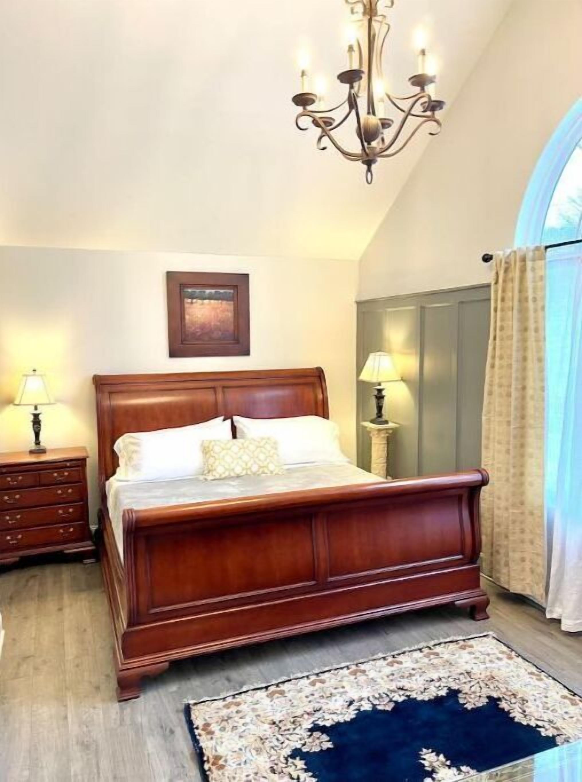 A bedroom with a wooden bed, a chandelier, a nightstand with a lamp, a dresser, a painting on the wall, and a view through a window with curtains.
