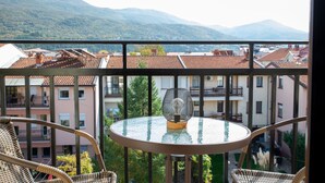Ein Balkon mit einem runden Glastisch und Korbstühlen, der auf ein Wohngebiet mit roten Ziegeldächern und einer Bergkulisse blickt.