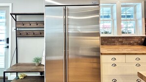 Un refrigerador de acero inoxidable en una cocina con piso de madera, un banco de madera y un ventanal con vistas a árboles.