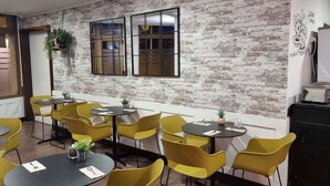 A room with yellow chairs and round tables, a brick wall, and a window with blinds. - HOTEL NORTE (Zarautz)