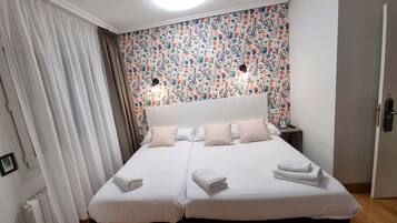 A double bed with white linens and two pillows, a nightstand with a lamp, and a colorful floral wallpaper.