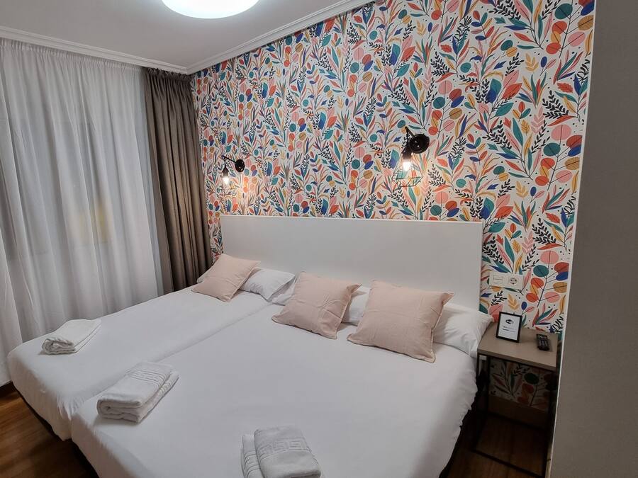 A neatly made bed with white and pink linens, a wall with a colorful leaf pattern, and a ceiling light.