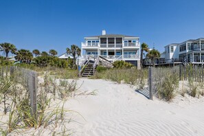 Une propriété située en bord de mer, comprenant un bâtiment à plusieurs niveaux, un escalier menant à l’entrée et une plage de sable agrémentée de végétation.