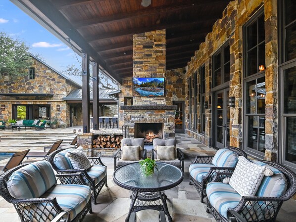 A covered outdoor patio with stone walls, a fireplace, wicker furniture, and a television screen.