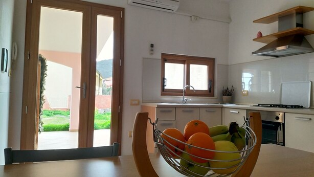 A modern kitchen with a dining area, featuring a large window, a built-in oven, and a fruit bowl on the table. - Panorama Apartment (Intimate, up to 3 people) (Muravera)