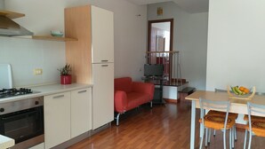 A compact kitchen with a gas stove, a microwave, and a small refrigerator. There is a red sofa and a dining table with chairs in the living area. A television is mounted on the wall. - Panorama Apartment (Intimate, up to 3 people) (Muravera)