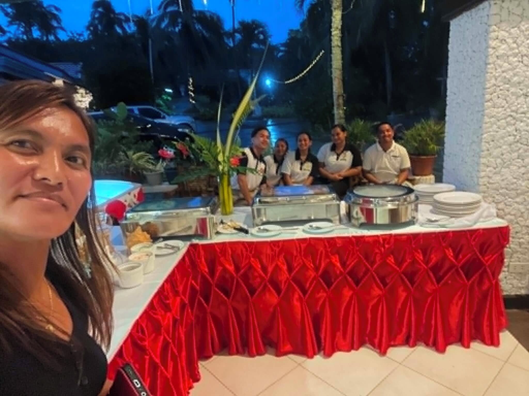 Un grupo de personas sentadas frente a una mesa de buffet con mantel rojo, rodeadas de diversos alimentos y platos.