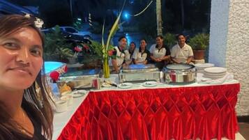 Un grupo de personas sentadas frente a una mesa de buffet con mantel rojo, rodeadas de diversos alimentos y platos.