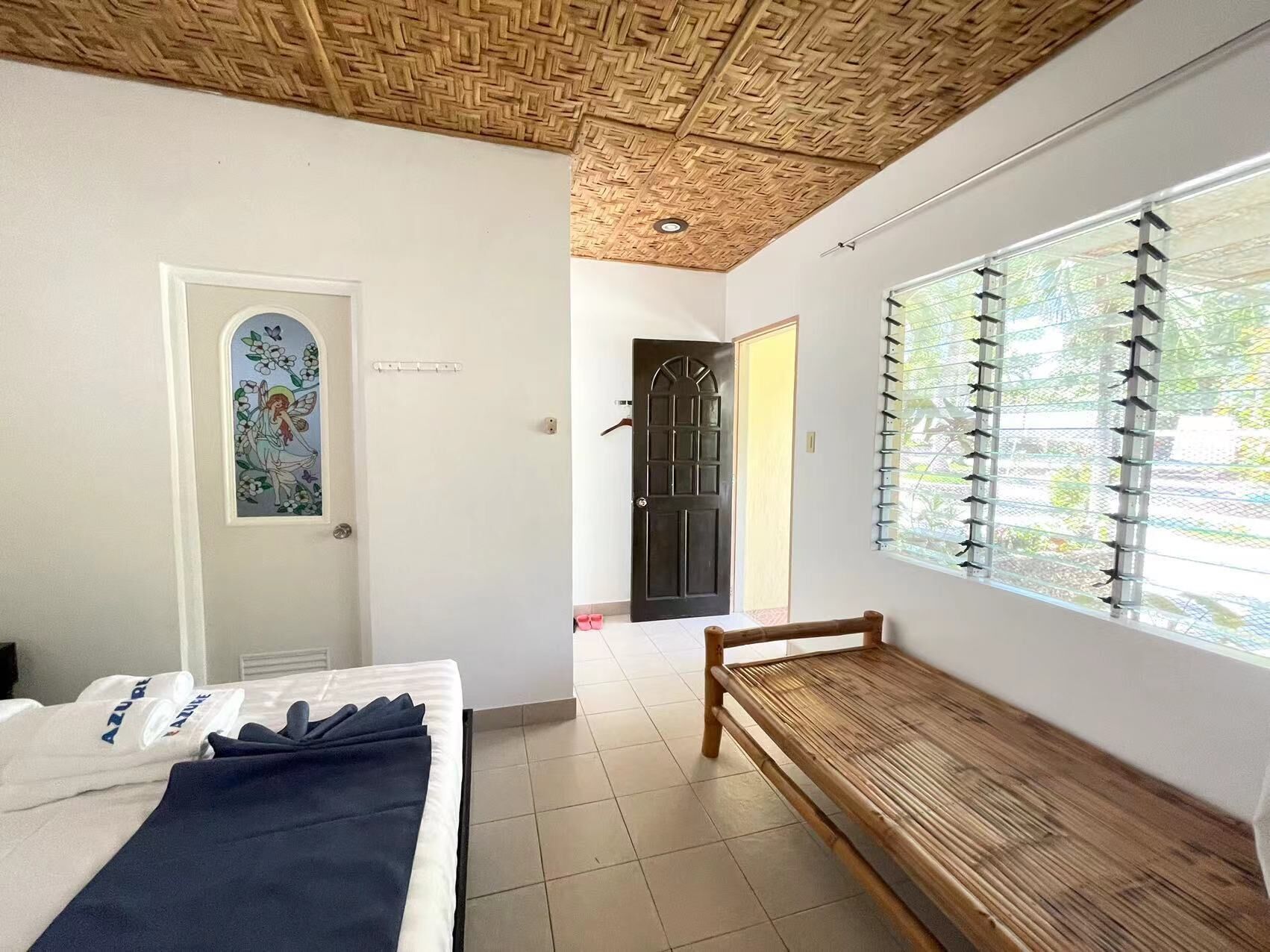 A room with a bed, a bench, a door with a decorative window, and a large window with blinds.