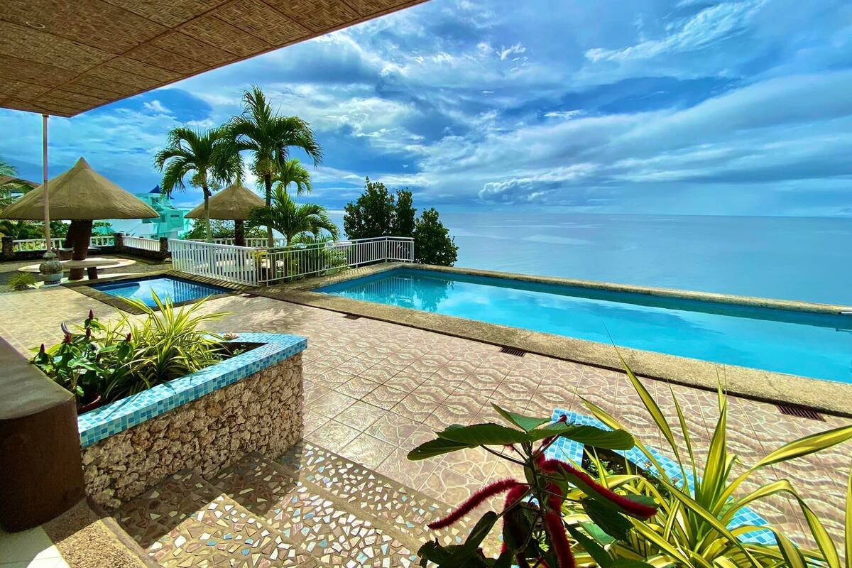 A pool area with a view of the sea, surrounded by palm trees and a thatched roof structure.