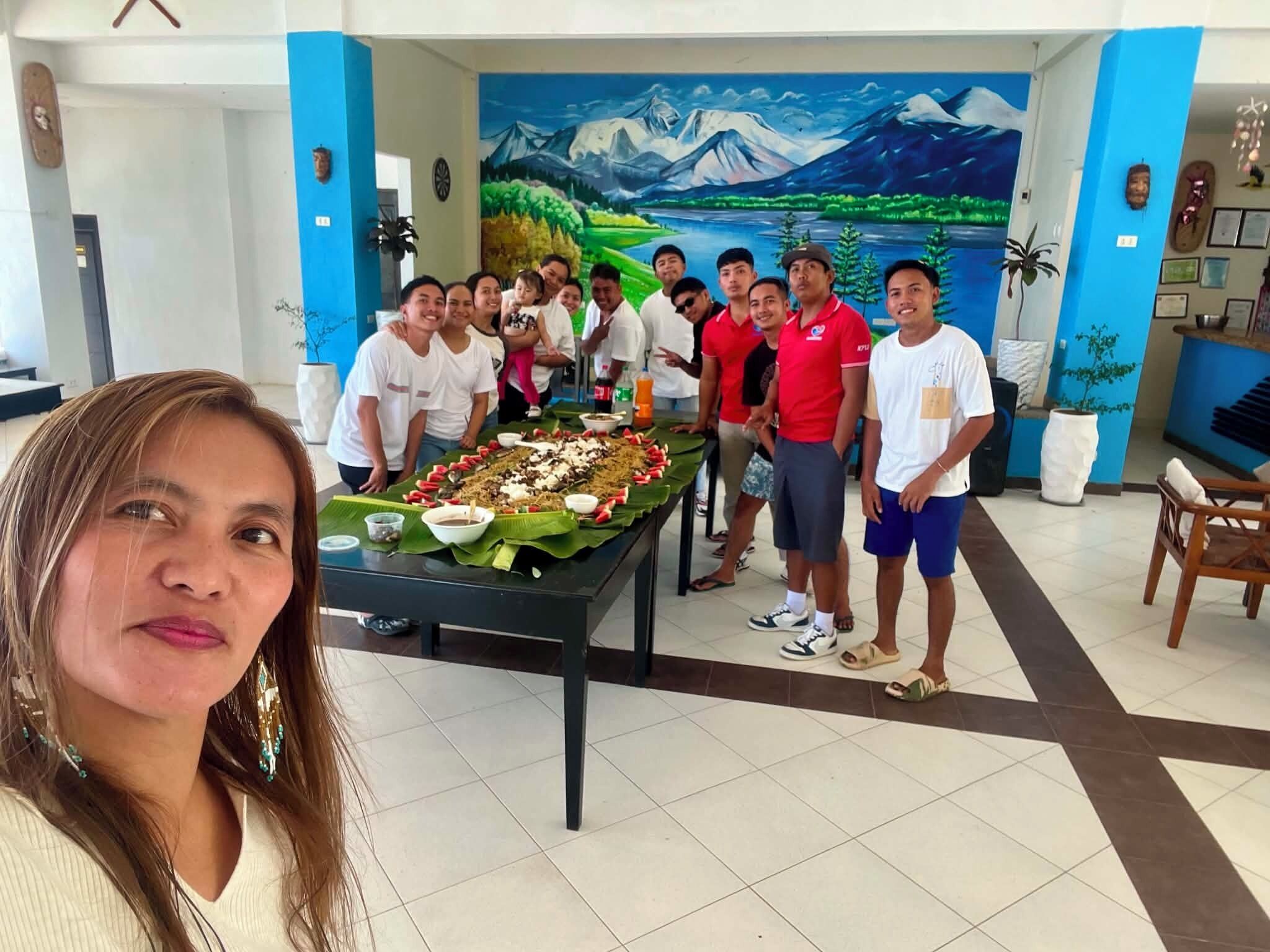A group of people gathered around a table with a meal, in a room with a mural of a mountainous landscape.