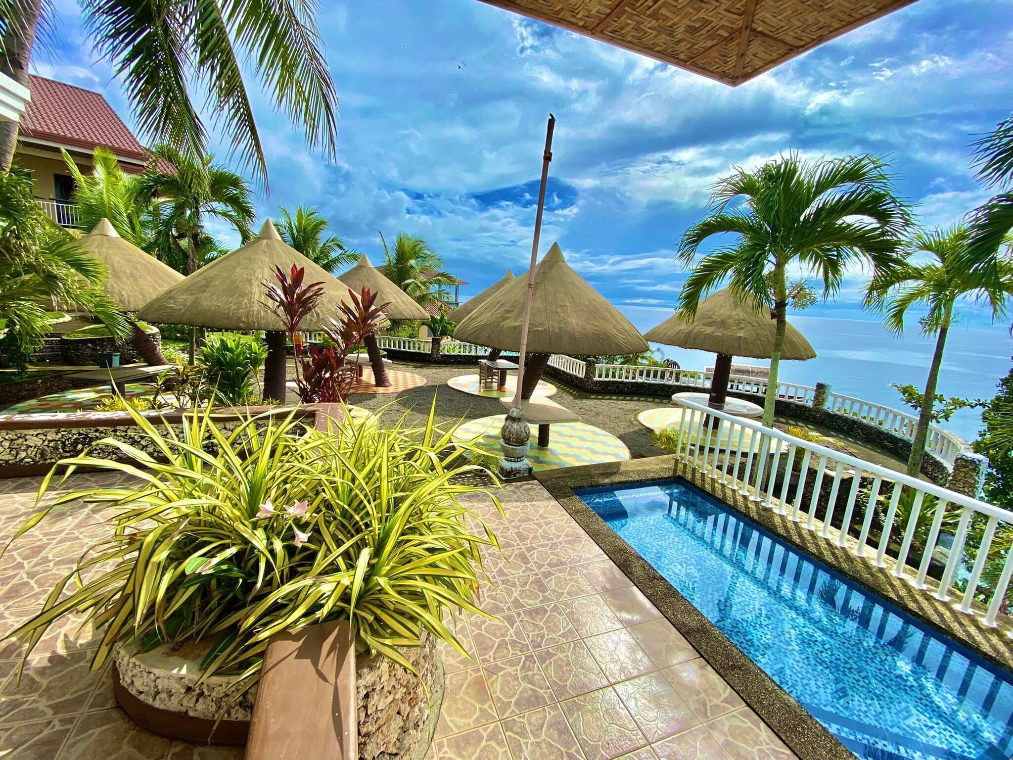 A pool area with a view of the ocean, surrounded by palm trees and thatched umbrellas.