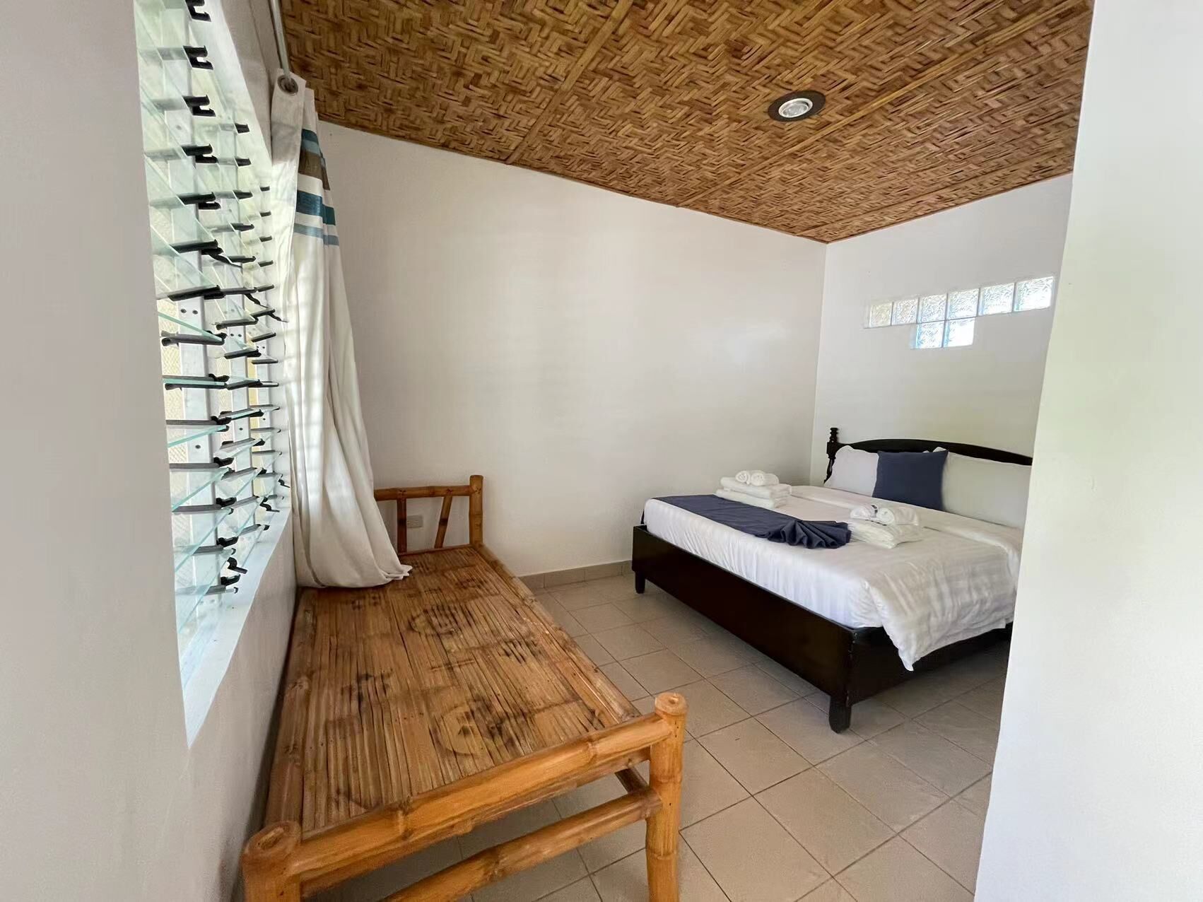 A room with a bed, a wooden bench, a wall-mounted shelf with glasses, and a woven ceiling.
