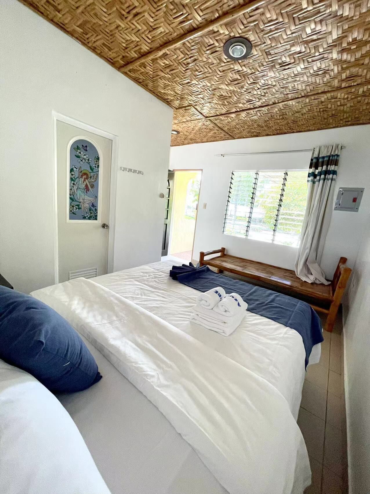 Ein Zimmer mit einem Bett, einer Bank, einer Tür mit dekorativer Verkleidung und einem Fenster mit Vorhängen.