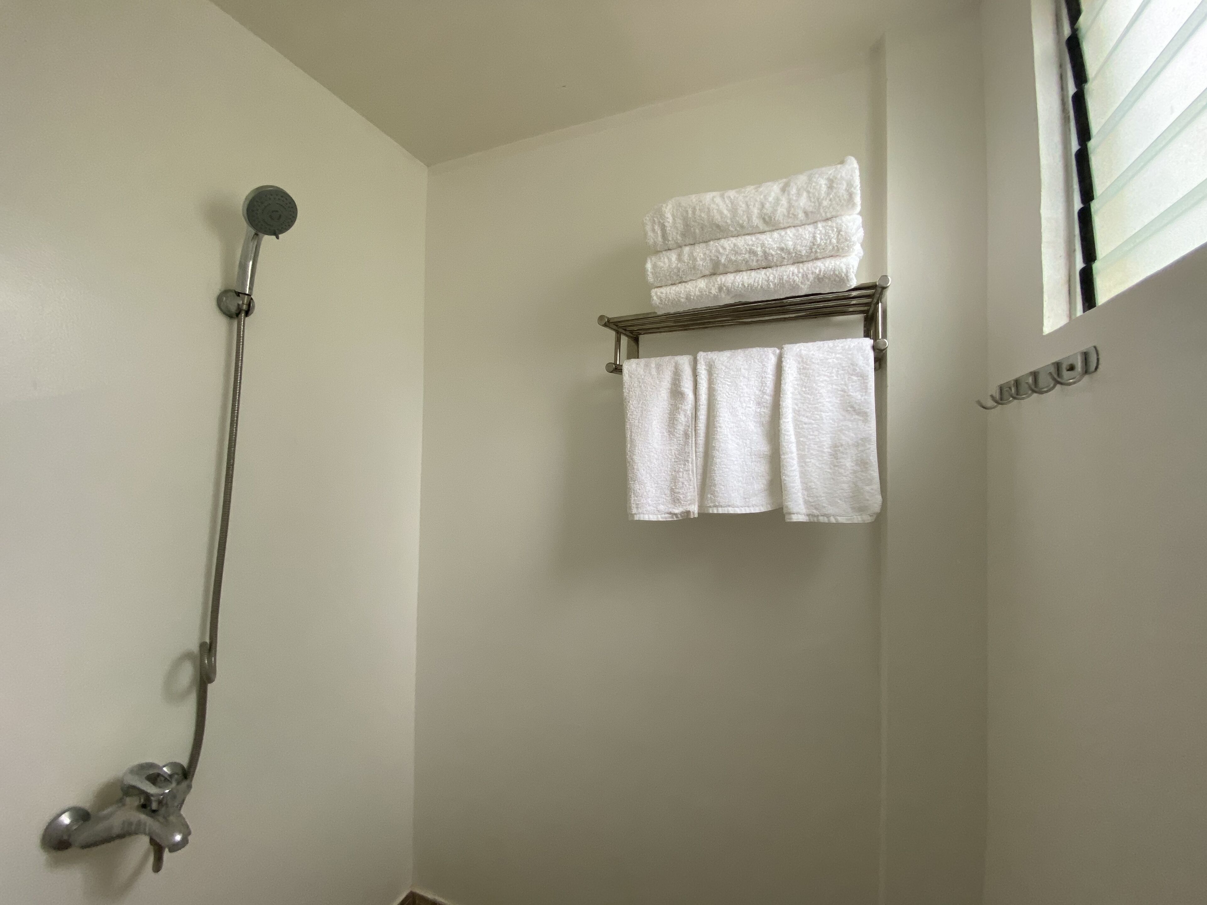 A bathroom with a shower, a towel rack with folded towels, and a window with blinds.