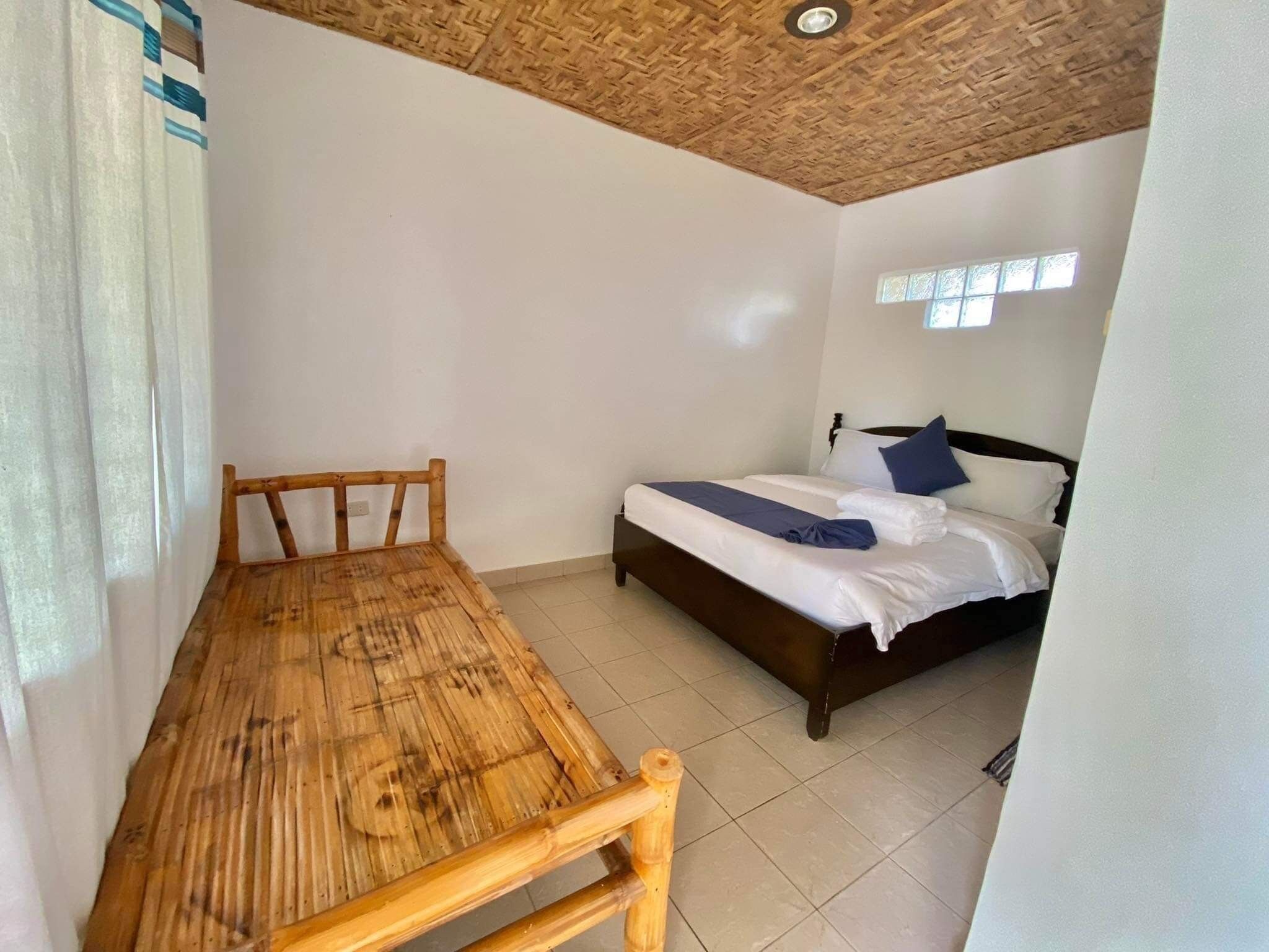 Een kamer met een houten tafel, een bed met wit beddengoed en blauwe kussens, en een raam met zonwering.