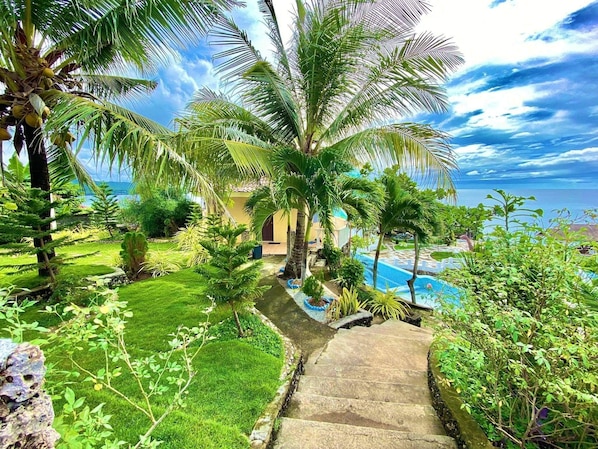 Un jardin tropical agrémenté de palmiers, une piscine et un escalier en pierre menant à un bâtiment.