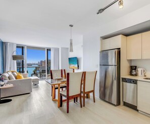 A modern apartment with a dining area, kitchen, and a large window overlooking a cityscape.