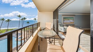 Un balcón con una mesa y sillas de vidrio, con vistas a una piscina y palmeras, y un cuadro en la pared.