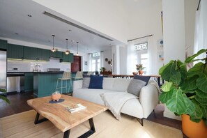 Eine moderne Küche mit grünen Schränken, ein Essbereich mit Holztisch und ein Wohnzimmer mit Sofa und großer Pflanze.