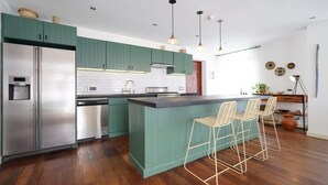 Une cuisine moderne avec des meubles de couleur verte, un réfrigérateur en acier inoxydable, un coin bar avec des tabourets et un éclairage suspendu.