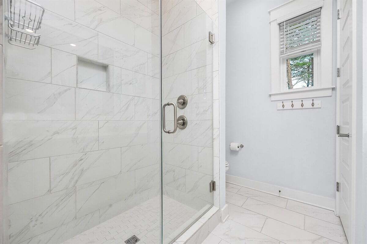 Une salle de bain dotée d’une douche à parois de verre, de carreaux de marbre blanc, d’une petite fenêtre avec des stores et d’un porte-serviettes.