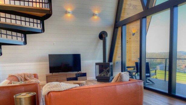 A modern living room with a large flat-screen TV, orange sofas, a fireplace, and a large window offering a view of the outdoors.
