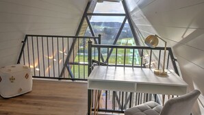 A room with a sloped ceiling, a white table, a lamp, a cushioned chair, and a view of a green landscape through a large window.