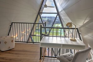 Huone, jossa on kalteva katto, valkoinen pöytä, lamppu, pehmustettu tuoli ja suuri ikkuna, josta avautuu näkymä vehreään maisemaan.