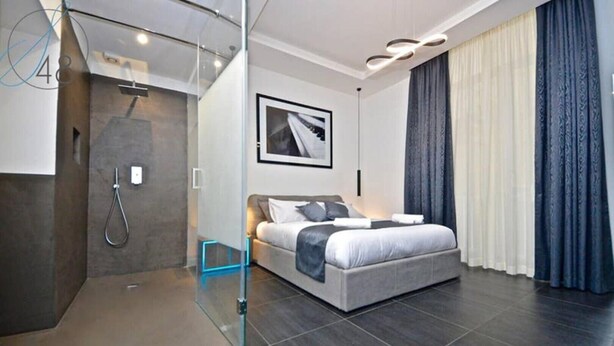 A modern hotel room with a glass-enclosed shower, a bed with grey bedding, and a large window with curtains. - Suitequarantotto CURS CIN IT063049C2Z4YC4Q9U  (Napoli)