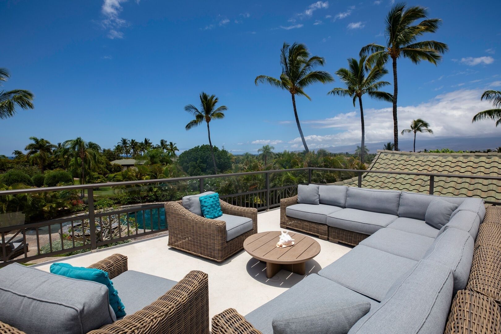A rooftop terrace with wicker furniture, a round table, and a view of palm trees and greenery.