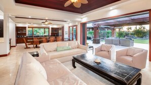 A spacious living room with a large sofa, a coffee table, and a dining area with chairs and a kitchen in the background. - Beach Walker: Exclusive w/ Pool,Views,Mauna Lani Beach,Golf/Pickleball (Kamuela)