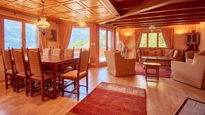 A spacious living area with a wooden dining table, chairs, and a sofa. There are large windows providing a view of the outdoors. - Family chalet overlooking the Midi Teeth (Champéry)