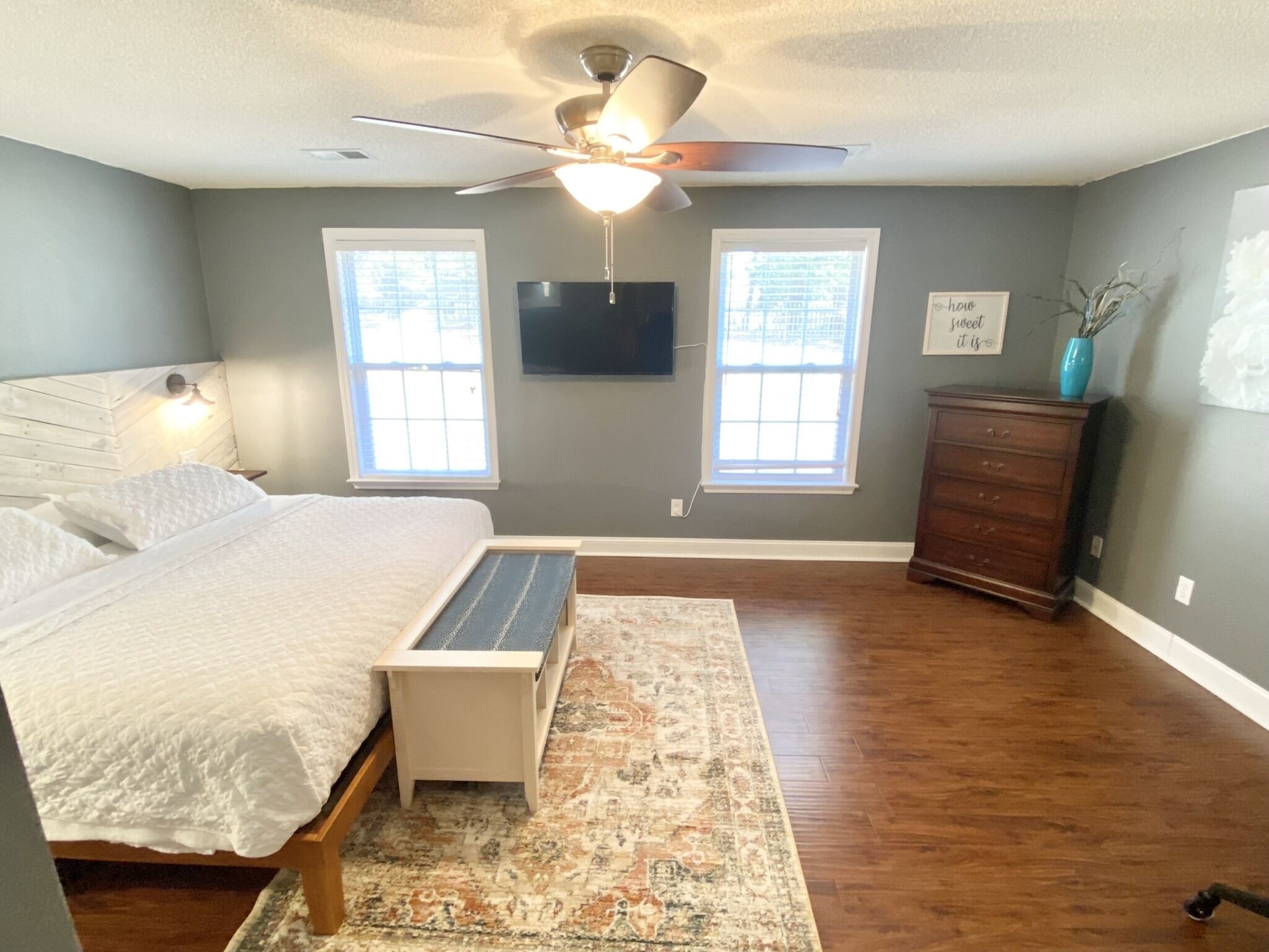 A bedroom with a bed, a desk, a dresser, a ceiling fan, and two windows.